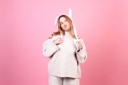 A beautiful girl in rabbit ears on a pink background grimaces, Easter holidays. The woman grimaces, kiss. Looks to the side, space for text.の写真素材