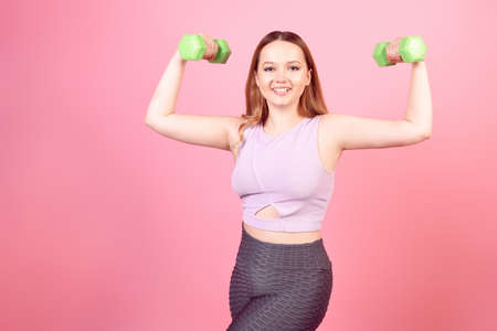 A beautiful girl goes in for sports on a pink background. The girl holds weights and shows exercises on her hands, sports goods, a healthy lifestyle. Woman in leggings.の写真素材