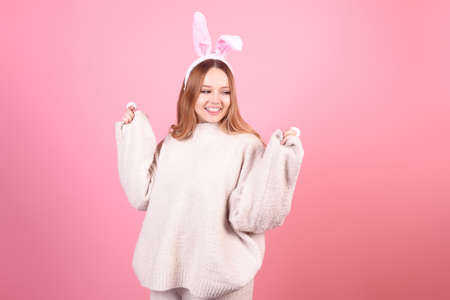 A girl with rabbit ears dances on a pink background. Easter holiday, smile and party. The girl is posing in the studioの写真素材