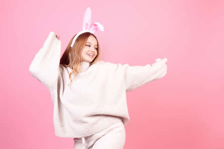 A girl with rabbit ears dances on a pink background. Easter holiday, smile and party. The girl is posing in the studioの写真素材