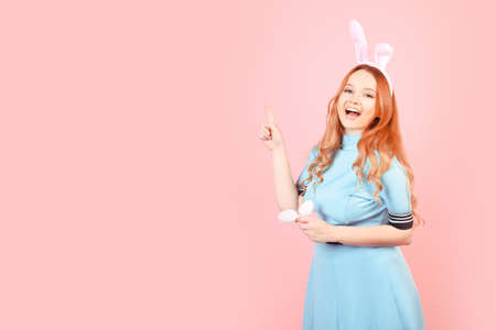 A girl with red hair in a blue dress and easter bunny ears on a pink background. A beautiful woman is pointing at a product or text.の写真素材