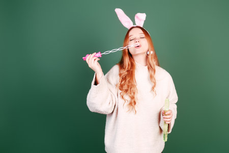 A beautiful red-haired girl with rabbit ears blows soap bubbles on a green background. Easter...