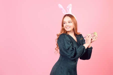 A beautiful girl in bunny ears paints Easter eggs on a pink background. Woman smiling and laughing,...