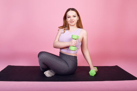 A beautiful body-positive girl on a pink background in a tracksuit goes in for sports, weights for training, exercises, fitness sports, yoga matの写真素材