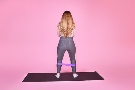 A beautiful body-positive girl on a pink background in a full-length sports suit goes in for sports, elastic bands for training, leg exercises, fitnessの写真素材