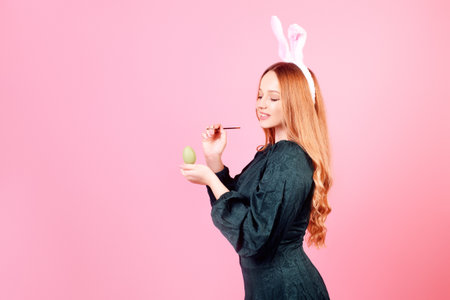 A beautiful girl in bunny ears paints Easter eggs on a pink background. Woman smiling and laughing, model with beautiful hair in green dressの写真素材