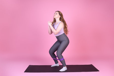 A beautiful body-positive girl on a pink background in a full-length sports suit goes in for sports, elastic bands for training, leg exercises, fitnessの写真素材