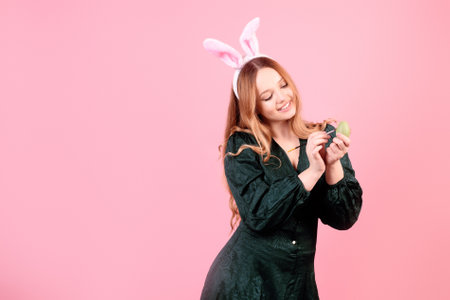 A beautiful girl in bunny ears paints Easter eggs on a pink background. Woman smiling and laughing, model with beautiful hair in green dressの写真素材