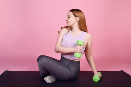 A beautiful body-positive girl on a pink background in a tracksuit goes in for sports, weights for training, exercises, fitness sports, yoga matの写真素材