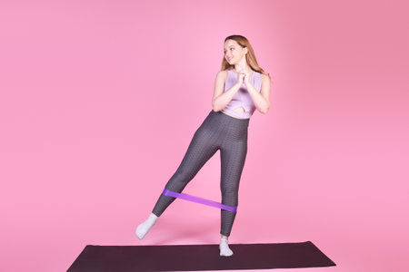 A beautiful body-positive girl on a pink background in a full-length sports suit goes in for sports, elastic bands for training, leg exercises, fitnessの写真素材