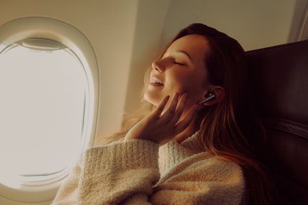 A beautiful girl is sitting in an airplane at the window listening to music or a podcast offline. Tourist travel.の写真素材