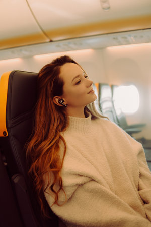 A beautiful girl is sitting in an airplane at the window listening to music or a podcast offline. Tourist travel.の写真素材