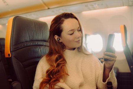 A beautiful brunette on an airplane looks at the phone and listens to music or watches a movie. Travel girl in flight.の写真素材