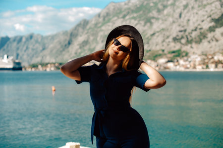 A woman in sunglasses and a hat stands against the backdrop of the sea and mountains, smiling. Vacation and outdoor recreation. Girl with long hair on vacation. Fashionの写真素材