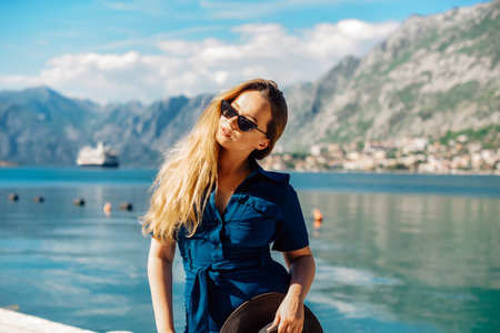 A woman in sun glasses and a hat stands against the backdrop of the sea and mountains. Vacation and outdoor recreation. Girl with long hair on vacationの写真素材