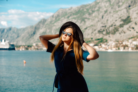 A woman in sun glasses and a hat stands against the backdrop of the sea and mountains. Vacation and outdoor recreation. Girl with long hair on vacation. Fashionの写真素材