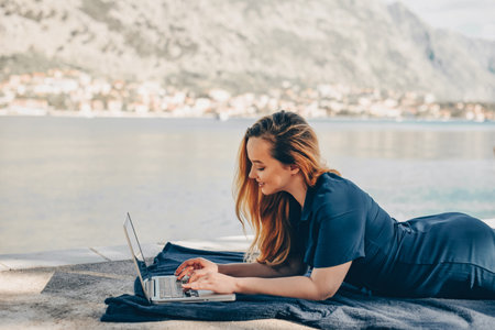 Girl freelancer lies on the background of the mountains with a laptop, remote work. A woman looks at the screen and writes something on the beach in the summer.の写真素材