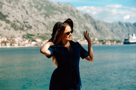 A woman in sunglasses and a hat stands against the backdrop of the sea and mountains, smiling. Vacation and outdoor recreation. Girl with long hair on vacation. Fashionの写真素材
