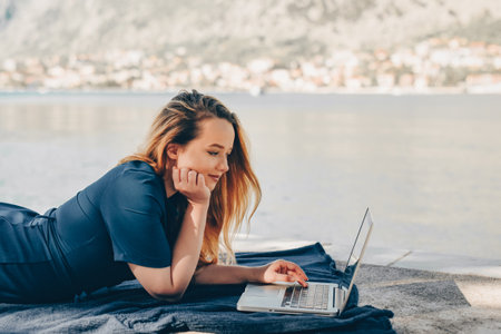 Girl freelancer lies on the background of the mountains with a laptop, remote work. A woman looks at the screen and writes something on the beach in the summer.の写真素材
