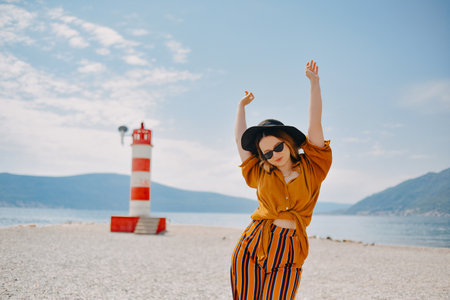 A girl in a yellow-orange suit on the beach against the background of the sea, mountains and a lighthouse. Fashion woman in sunglasses and hat dancingの写真素材