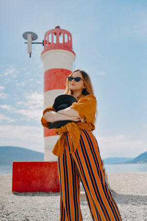 Fashionable woman on vacation against the backdrop of the sea, mountains and a lighthouse. Summer, sunny day, walk along the beach, enjoy life and travelの写真素材
