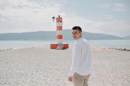 A man in a white shirt walks along the beach. The guy against the backdrop of mountains and the sea goes to the red lighthouse. Rest and travelの写真素材