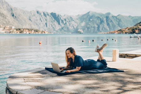 Girl freelancer lies on the background of the mountains with a laptop, remote work. A woman looks at the screen and writes something on the beach in the summer.の写真素材