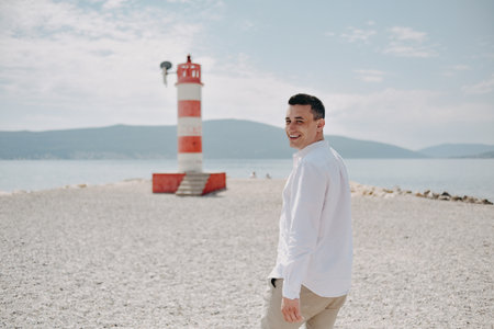 A man in a white shirt walks along the beach. The guy against the backdrop of mountains and the sea goes to the red lighthouse. Rest and travelの写真素材