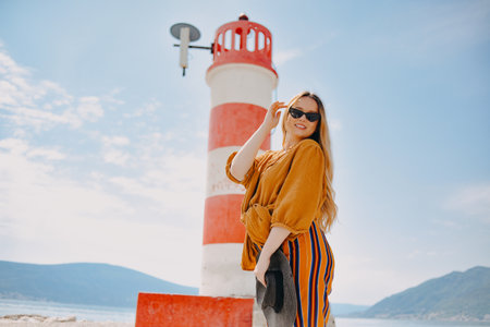 Fashionable woman on vacation against the backdrop of the sea, mountains and a lighthouse. Summer, sunny day, walk along the beach, enjoy life and travelの写真素材