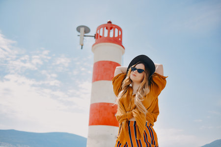 A girl in a yellow-orange suit on the beach against the background of the sea, mountains and a lighthouse. Fashionable woman in sunglasses and hat posingの写真素材