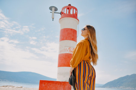 Fashionable woman on vacation against the backdrop of the sea, mountains and a lighthouse. Summer, sunny day, walk along the beach, enjoy life and travelの写真素材