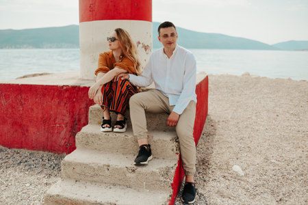 A couple sits near a lighthouse on the beach against the backdrop of the sea and mountains. Vacation love, honeymoon, travel. stylish coupleの写真素材