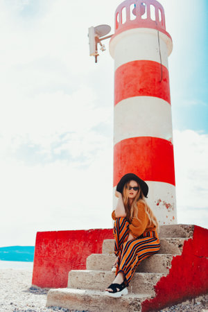 Gorgeous fashionable woman posing while sitting on a lighthouse. Summer photo of a girl in a yellow-orange suit, black hat and sunglasses. Blonde girl posing in travelの写真素材