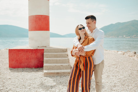 A couple stands near a lighthouse on the beach against the backdrop of the sea and mountains. Vacation love, honeymoon, travel. Stylish couple laughing and smilingの写真素材