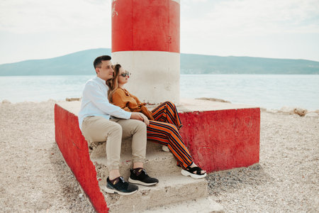A couple sits near a lighthouse on the beach against the backdrop of the sea and mountains. Vacation love, honeymoon, travel. stylish coupleの写真素材