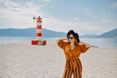 A girl in a yellow-orange suit on the beach against the background of the sea, mountains and a lighthouse. Fashionable woman in sunglasses and hat posingの写真素材