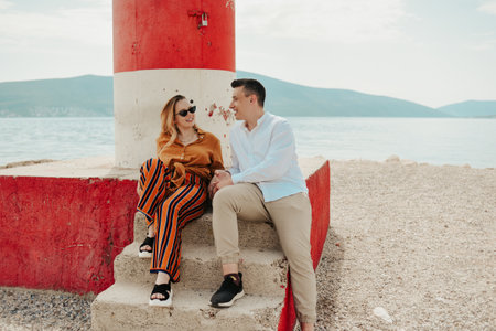 A couple sits near a lighthouse on the beach against the backdrop of the sea and mountains. Vacation love, honeymoon, travel. Stylish couple laughing and talking.の写真素材