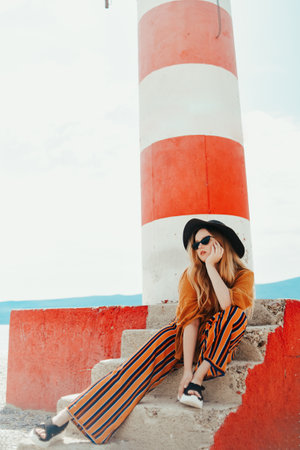 Gorgeous fashionable woman posing while sitting on a lighthouse. Summer photo of a girl in a yellow-orange suit, black hat and sunglasses. Blonde girl posing in travelの写真素材
