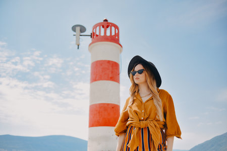 A girl in a yellow-orange suit on the beach against the background of the sea, mountains and a lighthouse. Fashionable woman in sunglasses and hat posingの写真素材
