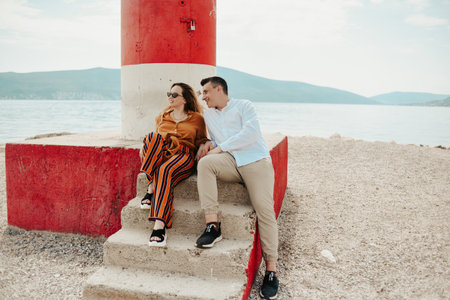 A couple sits near a lighthouse on the beach against the backdrop of the sea and mountains. Vacation love, honeymoon, travel. Stylish couple laughing and talking.の写真素材