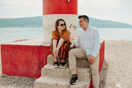 A couple sits near a lighthouse on the beach against the backdrop of the sea and mountains. Vacation love, honeymoon, travel. stylish coupleの写真素材