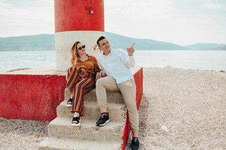 A couple sits near a lighthouse on the beach against the backdrop of the sea and mountains. Vacation love, honeymoon, travel. Stylish couple laughing and talking.の写真素材
