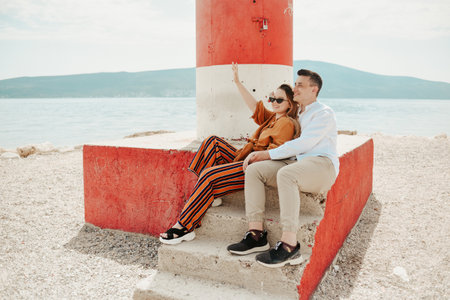 A couple sits near a lighthouseon the beach against the backdrop of the sea and mountains. Vacation love, honeymoon, travel. Stylish couple laughing and wavingの写真素材
