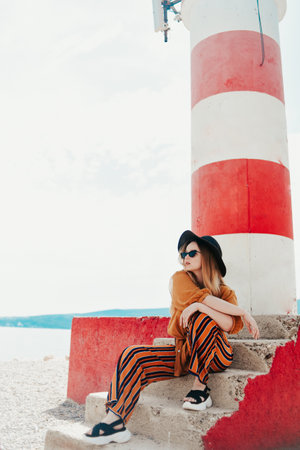 Gorgeous fashionable woman posing while sitting on a lighthouse. Summer photo of a girl in a yellow-orange suit, black hat and sunglasses. Blonde girl posing in travelの写真素材