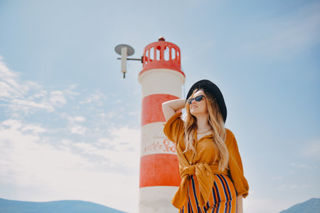 A girl in a yellow-orange suit on the beach against the background of the sea, mountains and a lighthouse. Fashionable woman in sunglasses and hat laughing and smilingの写真素材