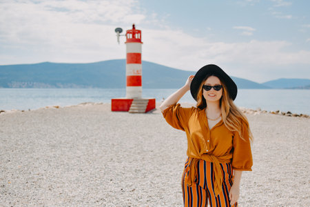 A girl in a yellow-orange suit on the beach against the background of the sea, mountains and a lighthouse. Fashionable woman in sunglasses and hat laughing and smilingの写真素材