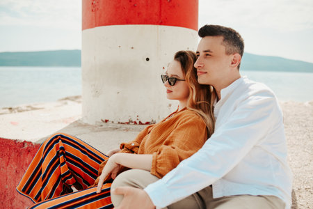 A couple sits near a lighthouse on the beach against the backdrop of the sea and mountains. Vacation love, honeymoon, travel. stylish coupleの写真素材