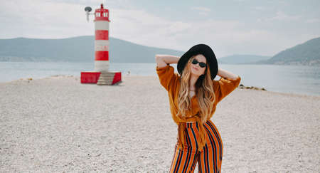 Fashionable woman on vacation against the backdrop of the sea, mountains and a lighthouse. Summer, sunny day, walk along the beach, enjoy life and travelの写真素材