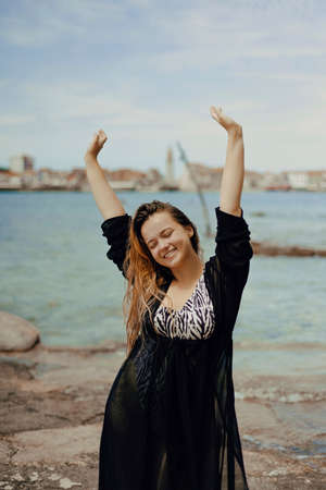 A gorgeous brunette on the beach against the backdrop of the sea and the European city smiles and laughs, enjoys her vacationの写真素材