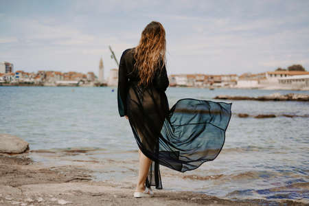 A woman with a flying pareo. Gorgeous brunette on the beach against the backdrop of the sea and the European city.の写真素材